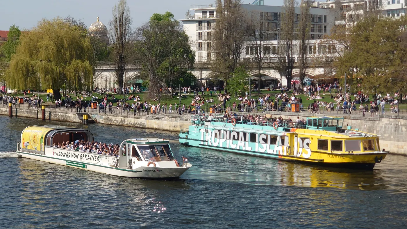 Zwei Touristenboote auf der Spree in Berlin. Im Hintergrund erholen sich viele Menschen auf einer grünen Wiese.