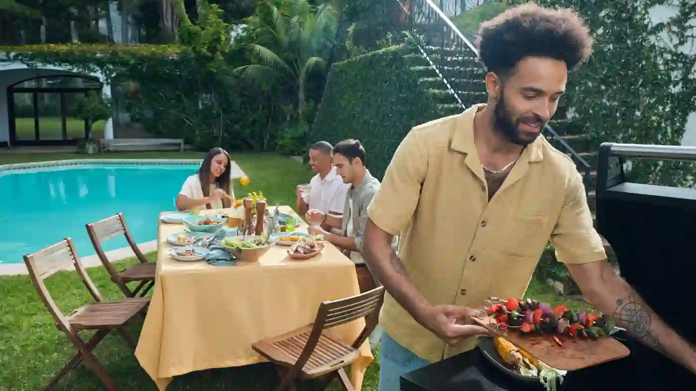 Ein Mann mit Afro-Frisur nimmt Gemüse-Spieße vom Grill. Im Hintergrund sitzen drei Personen im Garten an einem Tisch.
